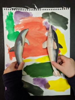  Aparecen Las manos de un niño llamado Alan sujetando dos figuras de Delfín uno más verde el otro más gris y los está poniendo de lado , verticalmente y en su fondo hay un papel pintado en colores con pinzeladas anchas de colores rojo, verde, amarillo lila, es una aportación de Alan & Beatriz